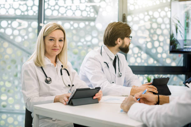 istock Successful smiling female Caucasian blond middle aged lady doctor, using tablet computer, smiling while sitting in light office with her colleagues and having video online conference 1285249563