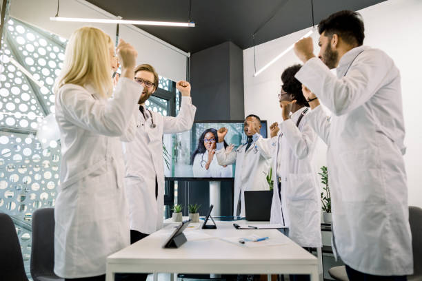 istock Medicine, conference, victory, successful trials. Five excited cheerful multiethnic doctors celebrating success, clenching fists and shouting, having video chat with their African female colleague 1285249612