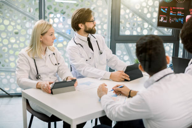 istock Cropped side angle view of multi-ethnic group of young doctors sitting around table during medical staff meeting or conference in clinic, and looking at the digital screen with charts and schemes 1285250299