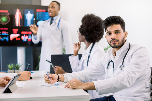 Portrait of young Indian male doctor, sitting at the table with his multiethnic colleagues, listening the speech of African man scientist, standing near big screen on the background.