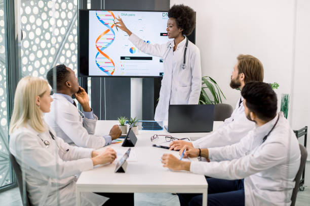 istock Group of multiethnical concentrated doctors in a conference during a training course, listening a speaker, young African woman scientist, talking about genetic engineering and pointing on the screen 1285250797
