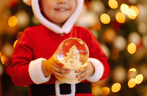Snow globe in the hand of Little child.  Kid in santa costume enjoys the snowfall. Winter holiday, New Year.