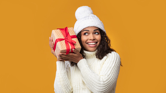 Guess What's Inside. Portrait Of Happy African American Woman Wearing Winter Hat Listening What Rattles In Gift Box. Curious Black Lady Shaking Christmas Present Isolated On Orange Background, Banner