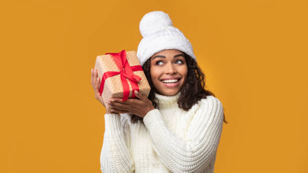 African American Woman Shaking Christmas Present At Studio Guess What's Inside. Portrait Of Happy African American Woman Wearing Winter Hat Listening What Rattles In Gift Box. Curious Black Lady Shaking Christmas Present Isolated On Orange Background, Banner christmas excitement stock pictures, royalty-free photos & images
