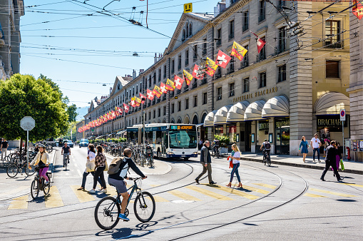 Geneva, Switzerland - September 4, 2020: Pedestrians and cyclists are crossing the rue de la Corraterie, with buildings decked with flags of Geneva and Switzerland, as a bus is waiting at a red light.