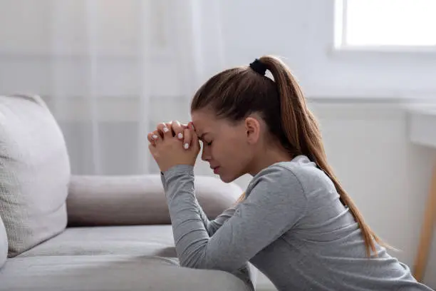 Side view of desperate young woman praying for help or consolation on sofa at home, copy space. Religious millennial lady worshiping God, asking for support in her grief or loss