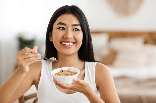 Happy asian woman holding bowl with homemade granola at bedroom. Healthy beautiful lady eating nutritive oatmeal at home, holding plate and spoon, smiling and looking at copy space, having breakfast