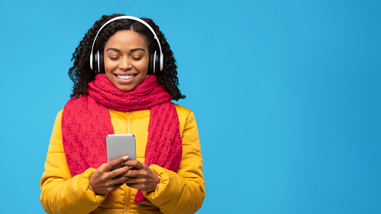 Young Black Woman Listening To Music On Phone Enjoying Favorite Song Standing Over Blue Studio Background, Wearing Warm Jacket. Musical Mobile App And Winter Playlist Concept. Panorama, Free Space