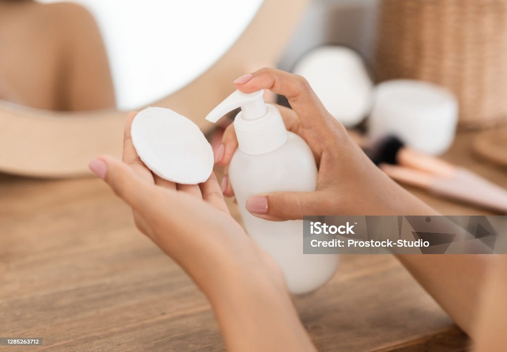 Unrecognizable woman applying beauty product on cotton pad Unrecognizable woman applying beauty product on cotton pad, standing next to mirror at home, cropped. Closeup of female hands holding cotton pad and bottle with toner or cleansing milk Facial Cleanser Stock Photo