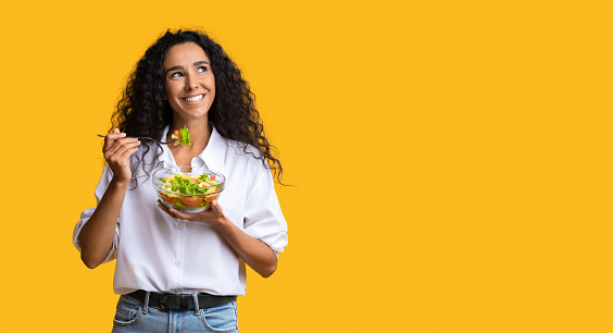 Cheerful Woman Eating Vegetable Salad From Bowl And Looking At Copy Space On Yellow Background, YoungLady Enjoying Heathy Nutrition And Organic Food, Having Vegetarian Meal For Lunch, Panorama