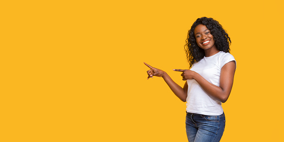 Cheerful black lady in white t-shirt pointing at copy space on yellow studio background. Smiling african american young woman showing exciting advertisement or announcement, panorama with empty space