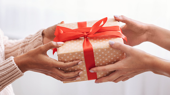 Holiday Celebration Concept. Closeup on female hands holding gift with red bow and giving present box to african american woman, greeting with Birthday, Christmas and New year at home