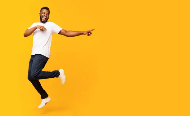 Joyful black millennial guy jumping up and pointing aside, panorama with free space, yellow studio background. Happy african american man jumping in the air, showing advertisement or text