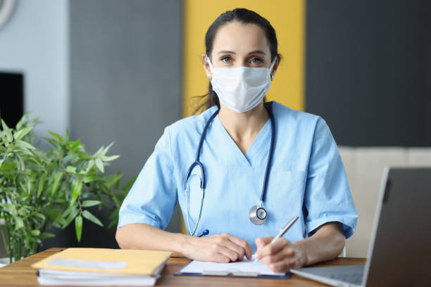 istock Doctor with protective medical mask siting at table in office and writing in documents 1285265908