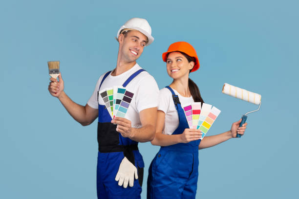 Cheerful Couple In Hard Hats Posing With Paint Brushes And Color Swatches Cheerful Couple In Hard Hats Posing With Paint Brushes And Color Swatches Over Blue Studio Background, Young Family In Work Clothes Choosing Palette To Repair Their Home, Ready For Renovation house painting service stock pictures, royalty-free photos & images