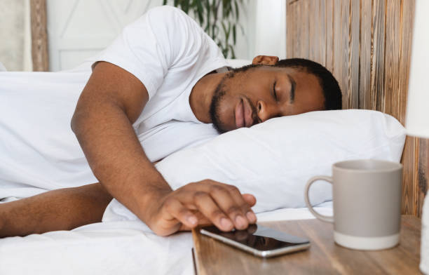 africano uomo dormire attraverso l'orologio di allarme su smartphone sdraiato nel letto - dormire troppo foto e immagini stock