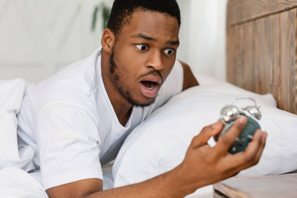 Shocked Black Guy Looking At Alarm-Clock Waking Up Oversleeping Indoors Oversleeping Concept. Shocked Black Guy Looking At Broken Alarm Clock Waking Up Late For Work In The Morning, Lying In Bed At Home. Awakening Problem and Tardiness, Bad Morning oversleeping stock pictures, royalty-free photos & images