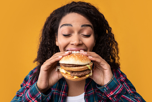 Gluttony. Closeup Portrait Of Funny Hungry African American Woman Eating Hamburger. Black Lady With Curly Hair Holding And Biting Tasty Fast Food, Isolated Over Orange Studio Background, Banner