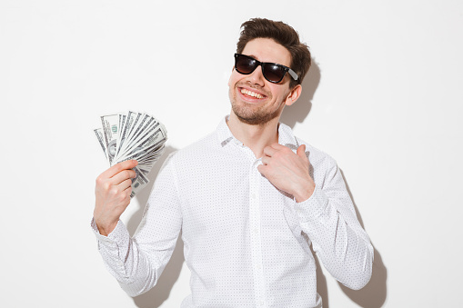 Portrait of a smiling young man in sunglasses holding money banknotes isolated over white background