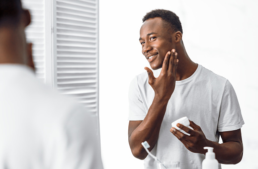 Handsome Black Man Using Facial Cream Moisturizing And Caring For Face Skin Standing Near Mirror In Bathroom Indoors In The Morning. Male Hygiene, Seflcare And Skincare Concept. Selective Focus