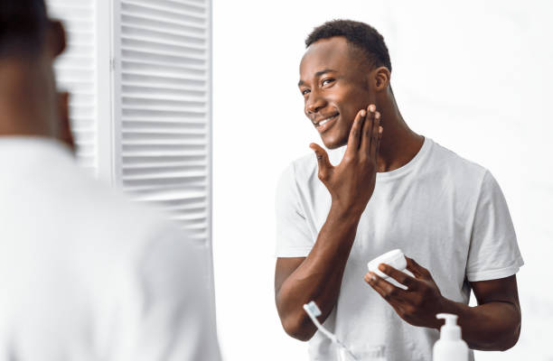 черный человек, используя крем для лица увлажняющий кожу лица в ванной комнате - male beauty male african descent ethnic стоковые фото и изображения