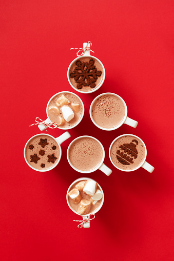 Cups with hot chocolate with marshmallows and cocoa powder decor in white mugs on red background stay Christmas tree shape, top view