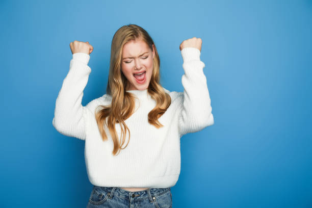 happy blonde beautiful woman in sweater showing yeah gesture isolated on blue background happy blonde beautiful woman in sweater showing yeah gesture isolated on blue background punching the air stock pictures, royalty-free photos & images