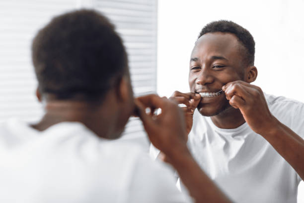 Toothy African Man Flossing Teeth Using Floss Standing In Bathroom Toothy African Man Flossing And Cleaning Teeth Using Tooth Floss Standing Near Mirror In Modern Bathroom At Home In The Morning. Toothcare And Healthy Oral Hygiene Concept. Selective Focus dental floss stock pictures, royalty-free photos & images