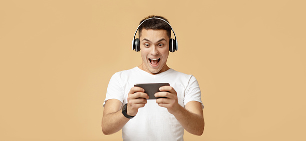 Cool game and online victory with modern gadget. Portrait of excited young man in headphones screams with joy and plays with smartphone, isolated on light background, studio shot, panorama, free space