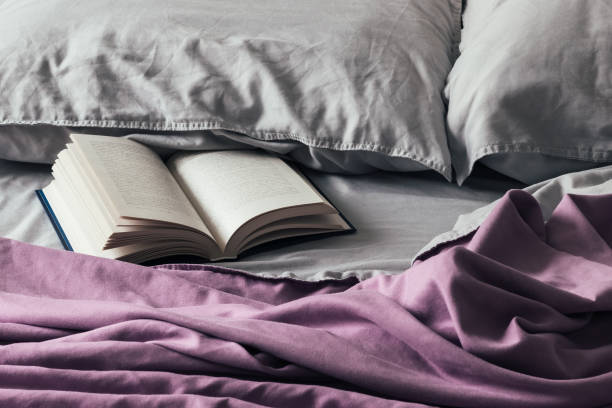 livre ouvert sur le lit en désordre avec un couvre-lit violet et des oreillers gris. livre fascinant dans la literie froissée douce - bedding cushion purple pillow photos et images de collection
