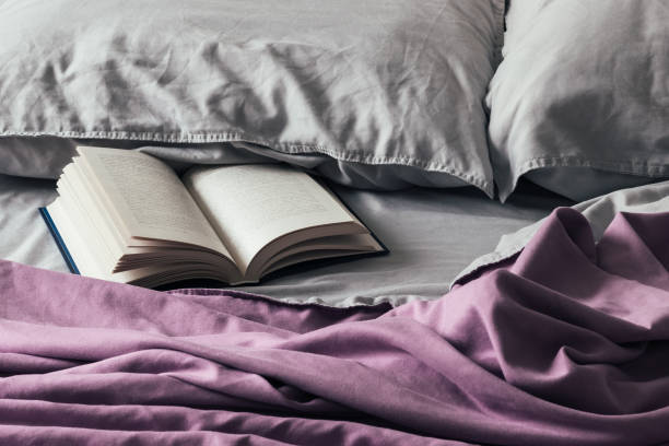 Open book on the messy bed with a purple bedspread and gray pillows. Fascinating book in soft crumpled bedding Open book on the messy bed with a purple bedspread and gray pillows. Fascinating book in soft crumpled bedding microfiber stock pictures, royalty-free photos & images