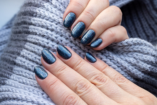 Female hands with gray knitted scarf with beautiful manicure - blue glittered nails. Selective focus. Closeup view