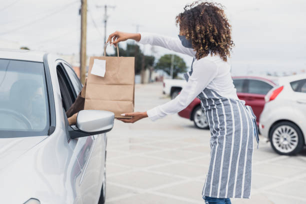Young adult waitress, wearing mask, social distances to deliver meal A young adult waitress wears her protective mask to deliver a curbside order to an unrecognizable person in a car.  The waitress stands away from the car. curbsidepickup stock pictures, royalty-free photos & images