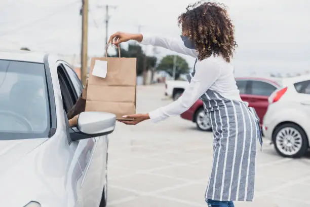 A young adult waitress wears her protective mask to deliver a curbside order to an unrecognizable person in a car.  The waitress stands away from the car.