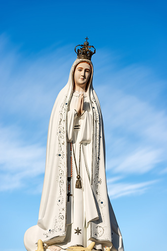 Closeup of a statue of Statue of Virgin Mary, mother of Jesus Christ, with crown and rosary on blue sky with clouds.