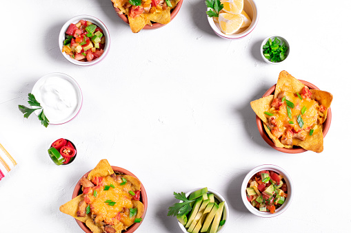 Nachos with chicken, mushroom and melted cheese in portion bowls served with assorted toppings on white table in a round. Salsa, avocado, sour cream, lemons, green onion. Copy space. Mexican food