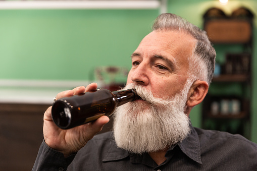 Older Caucasian bearded man, bearded model, in a retro hipster bar drinks a bottle of beer, photo portrait.