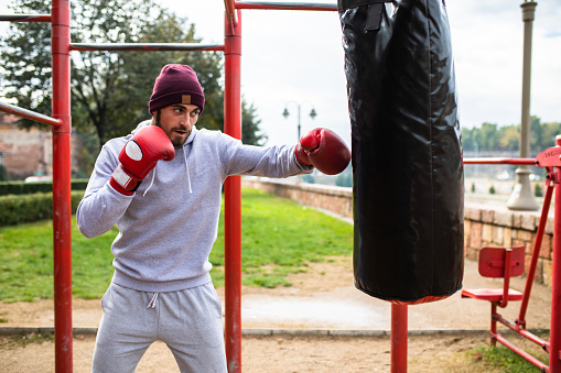 An attractive young fighter is punching a heavy bag in a street workout park on a rainy autumn day