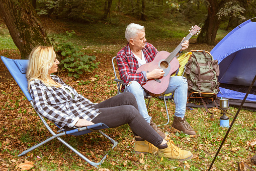 Beautiful middle aged couple enjoying on camping in nature. They play guitar next to the tent and campfire