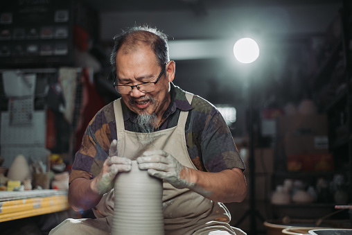 active senior man, owner of the pottery studio looking down with both hands making pottery