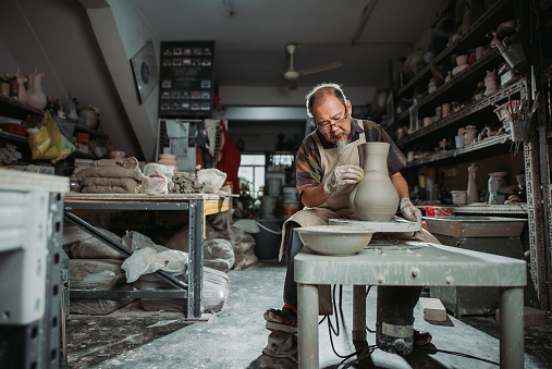 wide shot of active senior man, owner of the pottery studio looking down with hand making pottery