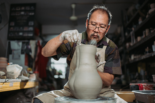 Asian chinese senior man clay artist making pottery on a spinning pottery wheel in his craft studio