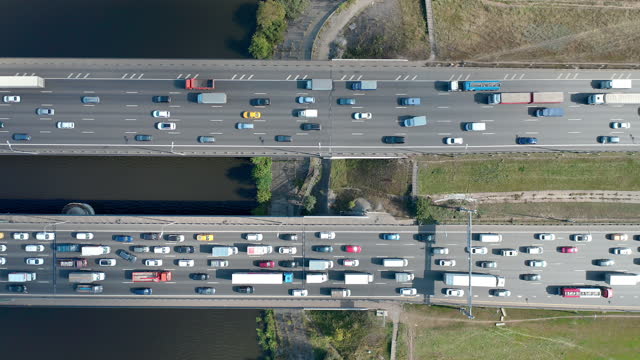 Traffic jam on the overpass over the coast. Double bridge with two separate roads. Big river under the bridge. Zoom out camera motion, moving up. Aerial top down view from rising drone. Sunlight weather. Summertime. High quality 4k footage.