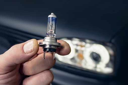 Car repair and service. The man holds in his hand a halogen light bulb against the background of the headlights on