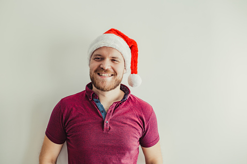 Young smiling man is wearing a Santa hat, he is standing in front of a white wall
