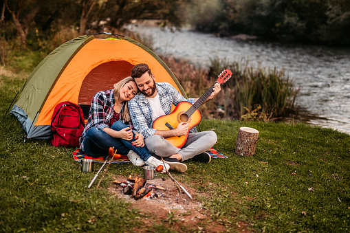 Cute heterosexual couple playing guitar and singing on camping by the river while grilling sausages on campfire.