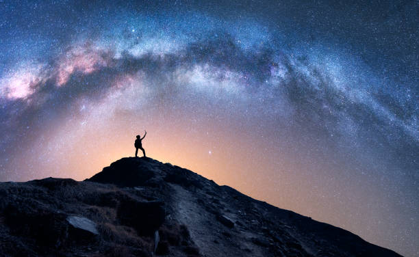 Photo of Arched Milky Way and happy man on the mountain at night. Silhouette of guy with raised up arm on the hill, sky with stars, yellow light in Nepal. Galaxy. Space landscape with milky way arch. Travel