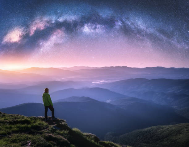 hombre en el pico de la montaña y arqueado vía láctea sobre montañas en nubes bajas por la noche. paisaje con cielo estrellado púrpura, arco de la vía láctea, luz naranja, hombre, colinas en la niebla. espacio y galaxia. cielo con estrellas - road street nature mountain peak fotografías e imágenes de stock