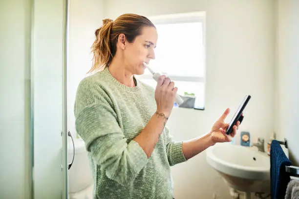 Woman standing in her bathroom at home brushing her teeth with an electric toothbrush and checking her cellphone