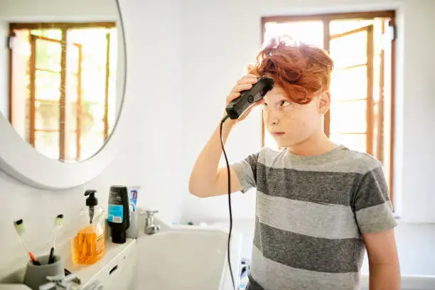 Young boy looking nervous while giving himself a trim with hair clippers in his bathroom at home